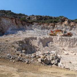 Das Bild zeigt einen Steinbruch mit einer großen, abgestuften Felswand und einem Bagger, der auf einer höheren Ebene steht. Im Vordergrund befindet sich eine breite, ausgehobene Fläche mit verstreuten großen Steinen und Erdmaterial. Der Himmel ist klar und blau, und die Umgebung wirkt trocken und felsig.