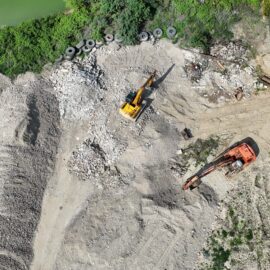 Das Bild zeigt eine Baustelle aus der Vogelperspektive mit zwei großen Baggern, die auf einer Fläche aus Schutt und Erde arbeiten. Neben den Maschinen liegen große Haufen aus Steinen und Baumaterial, während im oberen Bereich grüne Vegetation und ein kleiner Wasserbereich zu sehen sind. Mehrere alte Reifen sind am Rand der Baustelle abgelegt.