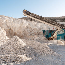 Das Bild zeigt eine große Maschine mit Förderband, die in einem Steinbruch arbeitet. Vor der Maschine liegen mehrere Haufen heller, grober Steine, während im Hintergrund eine hohe Felswand zu sehen ist. Die Szene ist von hellem Sonnenlicht beleuchtet und vermittelt den Eindruck intensiver Materialverarbeitung.