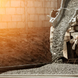 Das Bild zeigt einen Betonmischer, aus dessen Rutsche frischer Beton auf eine vorbereitete Fläche gegossen wird. Der Beton ist grobkörnig und verteilt sich auf einem Untergrund, der mit einer Metallarmierung versehen ist. Im Hintergrund sind eine Mauer aus hellen Betonblöcken und warmes Sonnenlicht zu sehen, das die Szene beleuchtet.