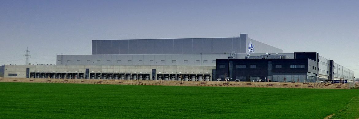 Ein großes Industriegebäude, vermutlich ein Lager- oder Logistikzentrum, mit mehreren Laderampen steht in offener Umgebung; im Vordergrund befindet sich eine grüne Wiese, darüber ein klarer Himmel.