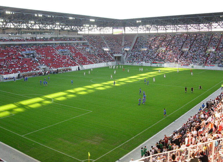 Fußballstadion mit roten Sitzplätzen, das größtenteils mit Zuschauern gefüllt ist; auf dem Spielfeld findet ein laufendes Spiel zwischen Mannschaften in blauen und weißen Trikots statt, mit Schattenwurf durch die Stadionüberdachung.