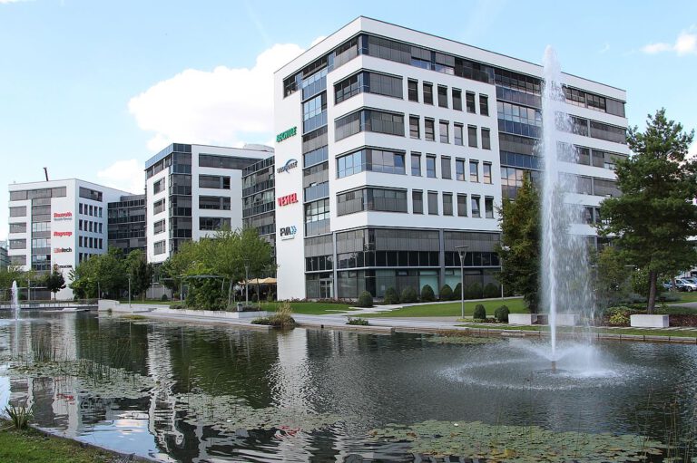 Ein modernes Bürokomplex mit mehreren mehrstöckigen Gebäuden umgibt einen großen Teich mit Springbrunnen, umgeben von Bäumen und Grünflächen; auf den Gebäuden sind Firmenlogos.