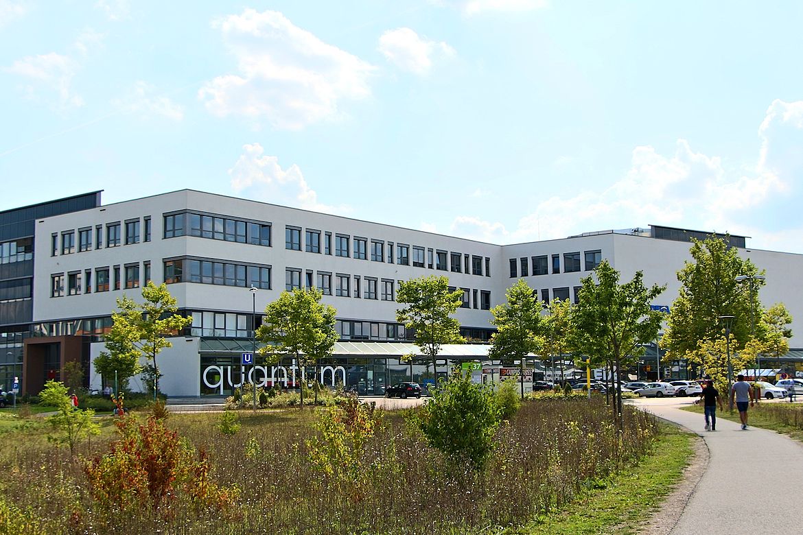 Modernes, mehrstöckiges Gebäude mit großen Fenstern und dem Schriftzug „quantum“ an der Fassade, umgeben von einer begrünten Anlage mit Gehweg, Bäumen und parkenden Autos unter teils bewölktem Himmel.
