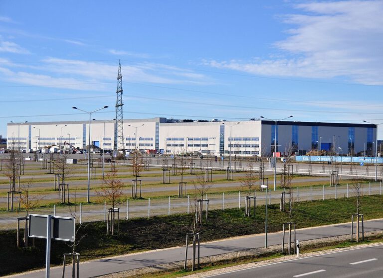 Ein großes Industrie- oder Lagergebäude mit mehreren Laderampen und Parkflächen steht hinter einer Reihe junger Bäume und Straßenlaternen entlang einer parallel verlaufenden Straße; im Hintergrund ist ein hoher Strommast unter einem überwiegend klaren Himmel mit einigen Wolken zu sehen.