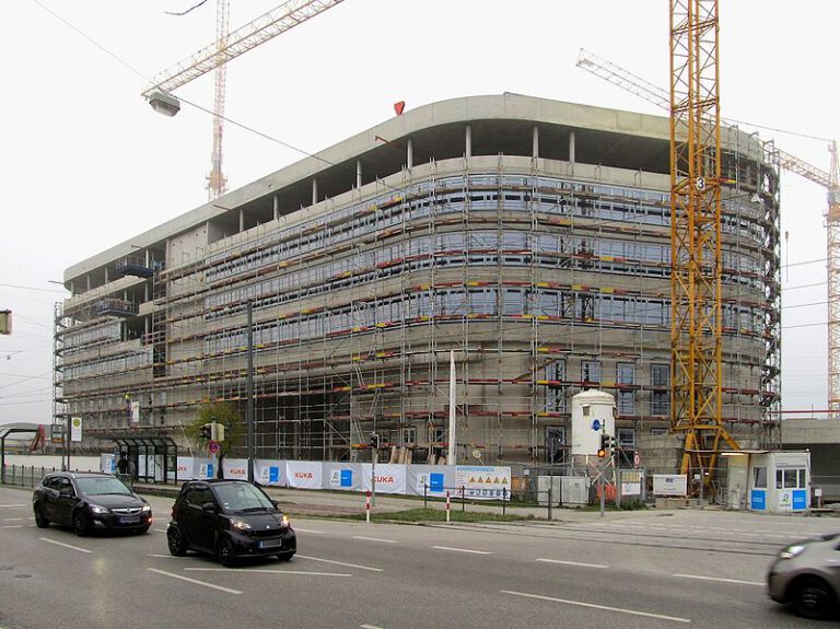 Ein großes Gebäude im Bau ist von Gerüsten umgeben und wird von mehreren Kränen umgeben; davor verläuft eine Straße mit vorbeifahrenden Autos sowie Baustellenschildern und Absperrungen entlang des Geländes.