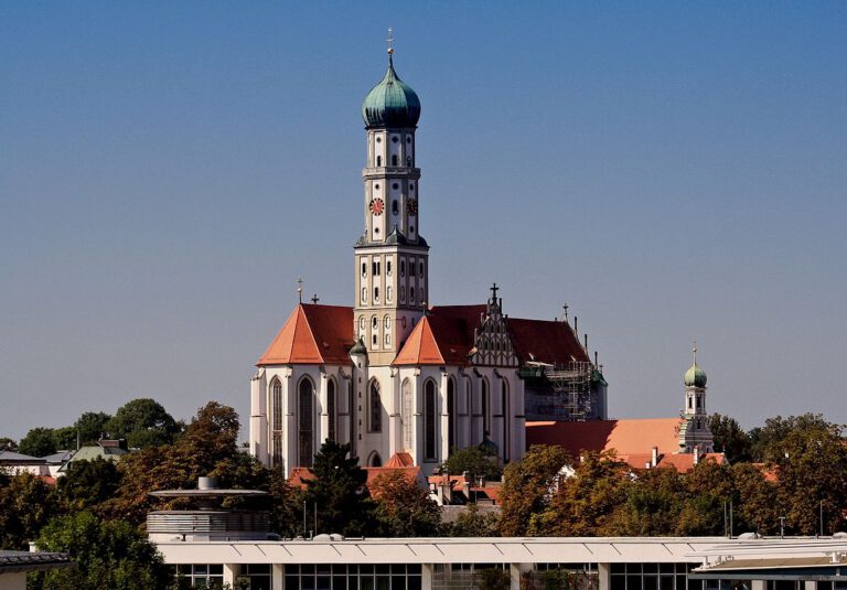 Historische Kirche mit hohem zentralen Turm und grünem Kuppeldach, umgeben von roten Ziegeldächern, Bäumen und weiteren Gebäuden.