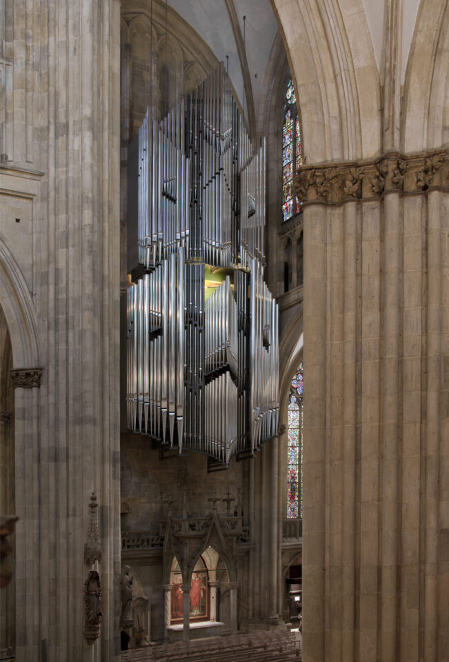 Innenraum einer großen Kathedrale mit hohen, verzierten Säulen und Bögen; im Mittelpunkt eine große Orgel mit zahlreichen Metallpfeifen, im Hintergrund bunte Glasfenster und architektonische Details.
