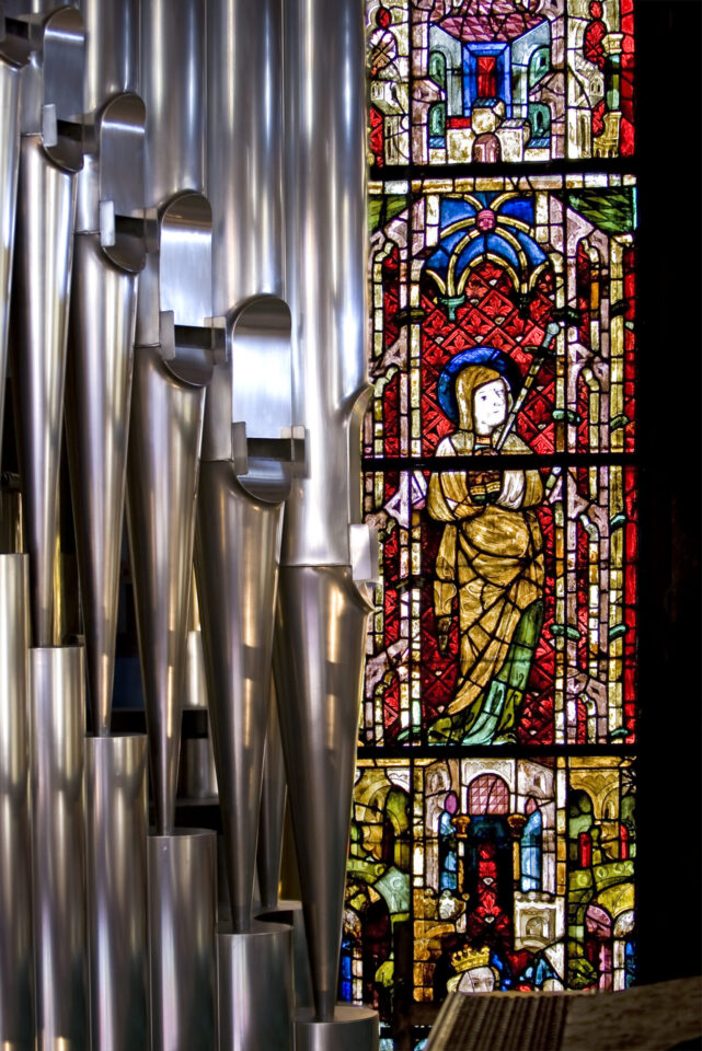 Kircheninnenraum mit großen metallischen Orgelpfeifen auf der linken Seite und einem farbigen Glasfenster mit einer religiösen Figur in Gewändern auf der rechten Seite.