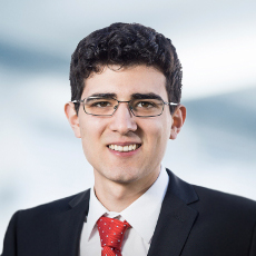 Das Bild zeigt eine Person in einem schwarzen Anzug mit weißem Hemd und roter Krawatte mit weißen Punkten. Der Hintergrund ist unscharf und zeigt blaue und weiße Farbtöne.