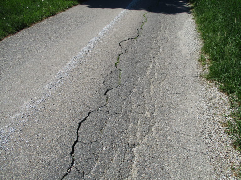 Das Bild zeigt eine Nahaufnahme einer rissigen und abgenutzten Asphaltstraße. Ein großer Riss verläuft längs durch die Fahrbahn, aus dem an einigen Stellen Gras wächst. In der Mitte ist eine weiße gestrichelte Linie zu sehen, und auf beiden Seiten der Straße befindet sich grünes Gras.