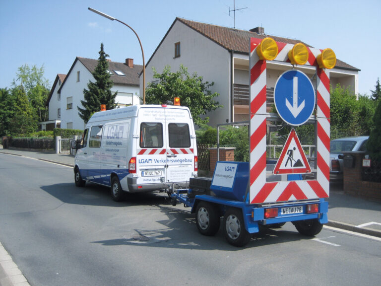 Das Bild zeigt ein Straßeninstandhaltungsfahrzeug mit einem Anhänger, auf dem ein großes Verkehrsschild montiert ist. Das Schild weist mit Leuchten und Symbolen auf Straßenbauarbeiten hin und gibt an, dass Fahrzeuge rechts vorbeifahren sollen. Im Hintergrund sind Wohnhäuser und Grünflächen zu sehen.