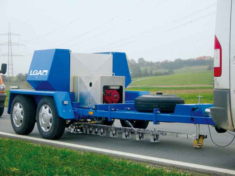 Das Bild zeigt einen blauen Anhänger mit zwei Rädern pro Seite, der an ein weißes Fahrzeug gekoppelt ist. Auf dem Anhänger befinden sich verschiedene Maschinen, darunter ein rotes Bauteil in einem Metallgehäuse sowie ein Ersatzrad. Die Ausrüstung unter dem Anhänger deutet auf eine Nutzung für Straßenwartung oder Prüfzwecke hin, im Hintergrund sind grüne Felder und Stromleitungen zu sehen.