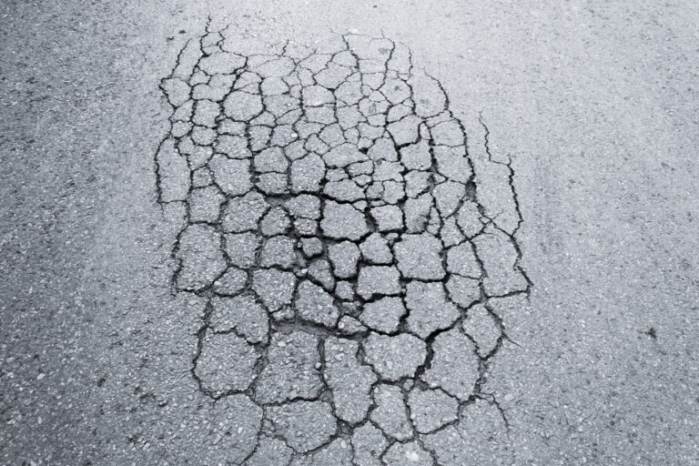 Das Bild zeigt eine stark beschädigte Asphaltfläche mit zahlreichen Rissen. Die Risse verlaufen unregelmäßig und deuten auf eine erhebliche Abnutzung der Straßenoberfläche hin.