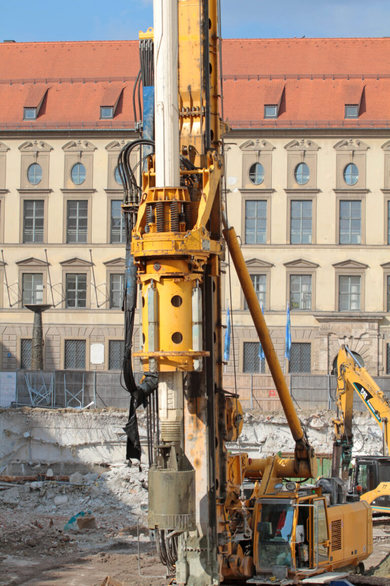 Das Bild zeigt eine Baustelle mit einer großen gelben Bohrmaschine im Vordergrund, die mit hydraulischen Komponenten und Kabeln ausgestattet ist. Im Hintergrund steht ein älteres Gebäude mit mehreren Fenstern und einem roten Ziegeldach. Auf dem Gelände sind außerdem weitere Baumaschinen und Bauschutt zu sehen.