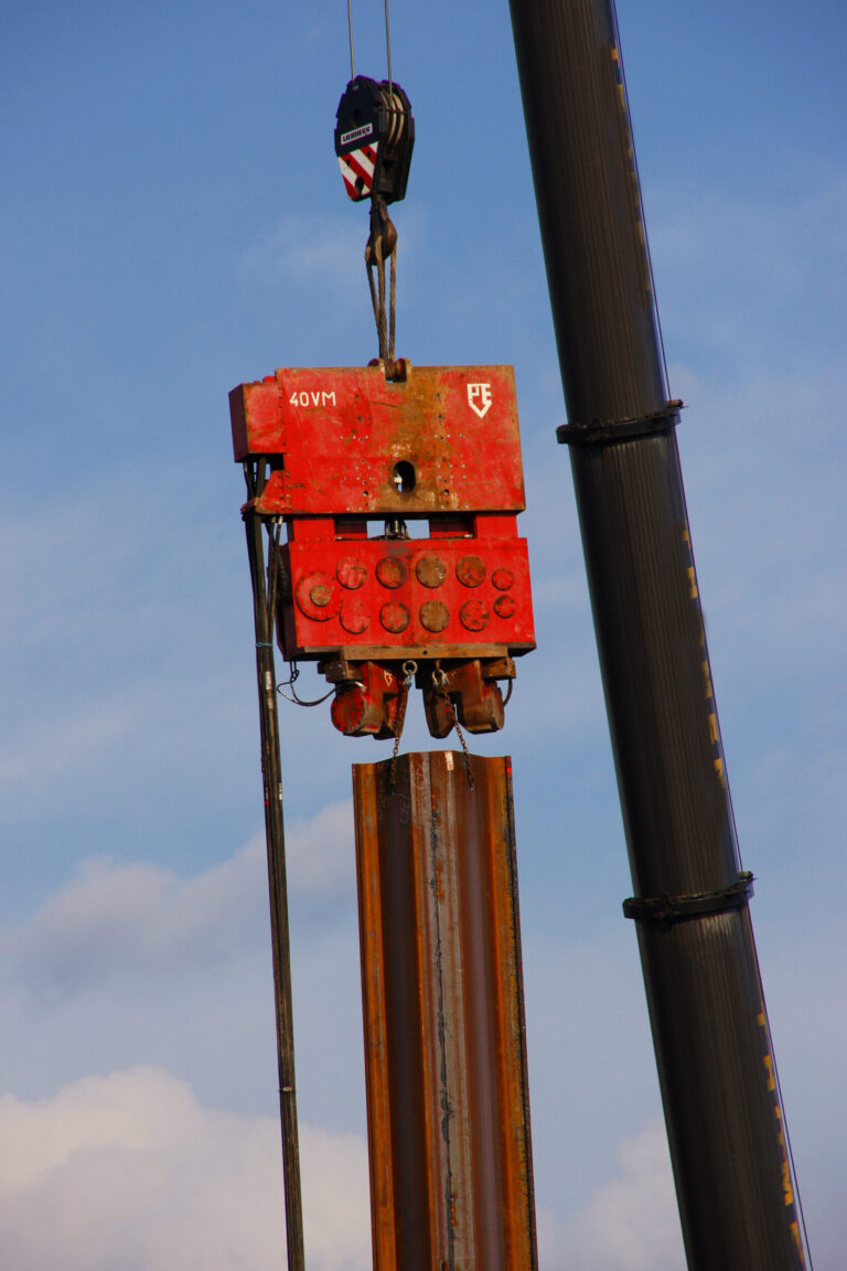 Das Bild zeigt eine Baustelle mit einem schweren roten Vibrationshammer, der an einem Kran befestigt ist. Das Gerät wird verwendet, um einen Stahlträgerpfahl in den Boden zu treiben oder herauszuziehen. Im Hintergrund ist ein klarer Himmel mit einigen Wolken zu sehen.