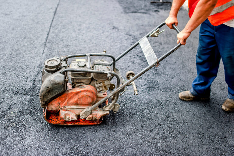 Das Bild zeigt eine Person, die mit einer Rüttelplatte frisch verlegten Asphalt verdichtet. Die Person trägt eine orangefarbene Warnweste und eine blaue Hose. Die Rüttelplatte ist orange und dient dazu, die Asphaltoberfläche gleichmäßig zu verdichten.