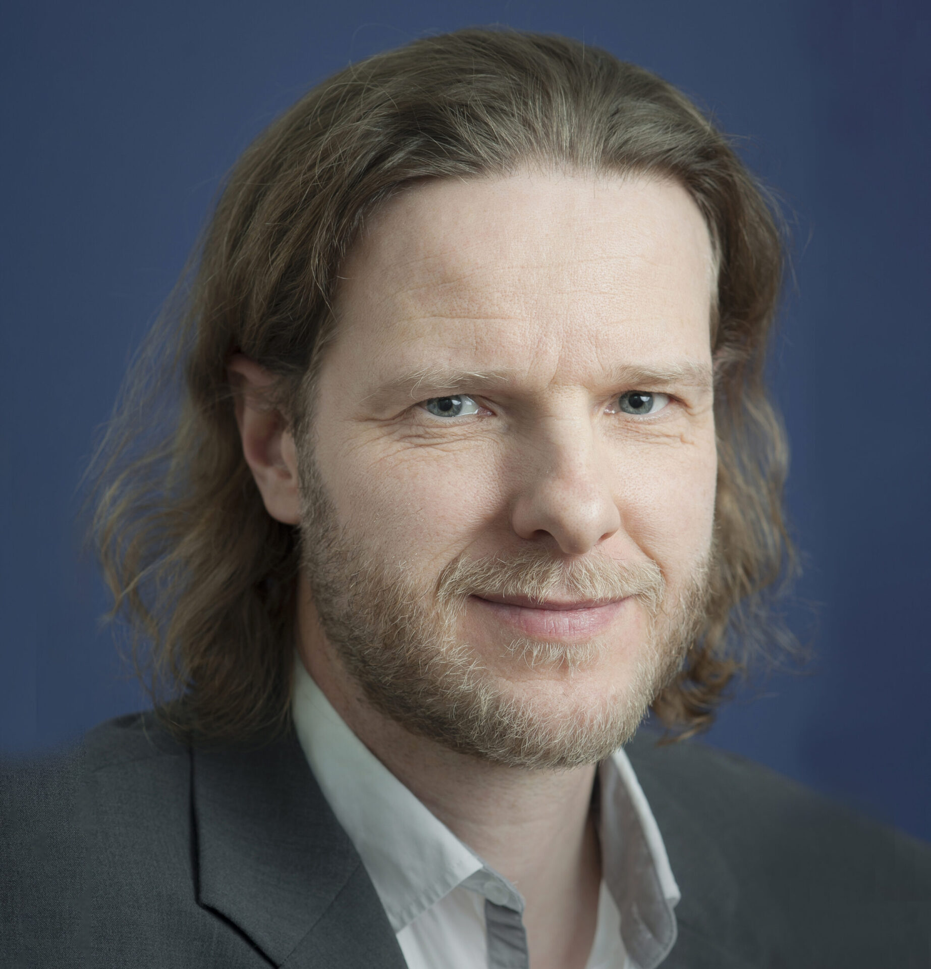 Ray Estel trägt ein Sakko und ein Hemd mit Kragen. Der Hintergrund ist unscharf und zeigt ein Büro.
