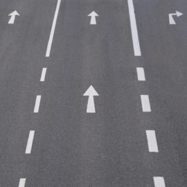 Das Bild zeigt eine mehrspurige Straße mit Fahrbahnmarkierungen und Richtungspfeilen. Es sind fünf Fahrspuren zu sehen, die jeweils durch weiße gestrichelte Linien und Pfeile gekennzeichnet sind. Die ersten vier Spuren erlauben die Geradeausfahrt, die fünfte Spur erlaubt sowohl das Abbiegen nach rechts als auch das Geradeausfahren.