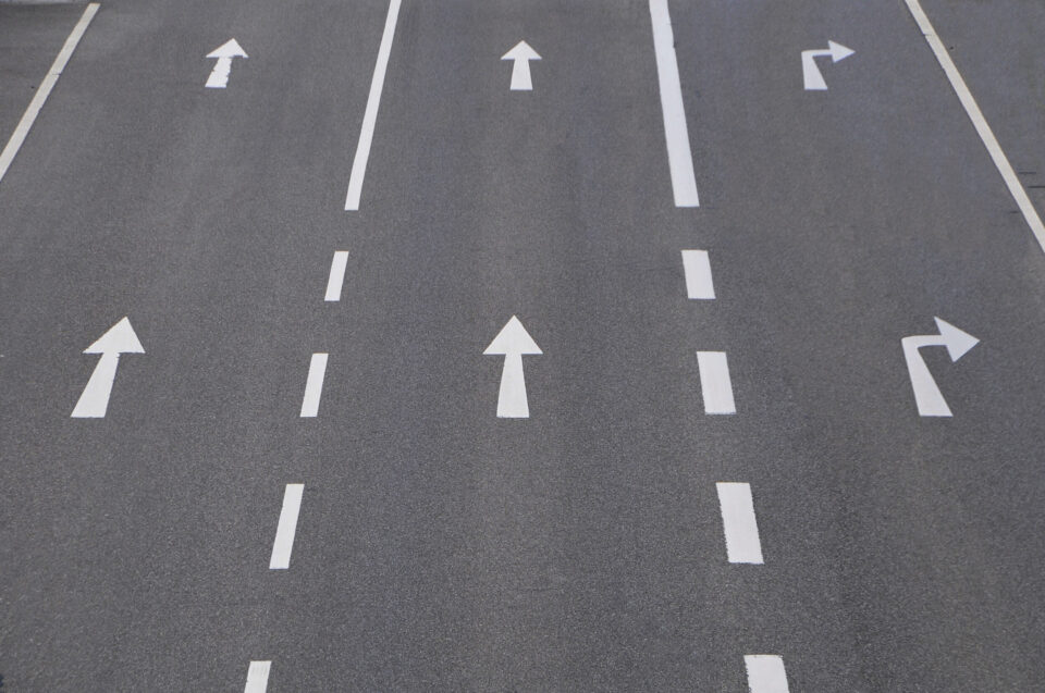 Das Bild zeigt eine mehrspurige Straße mit Fahrbahnmarkierungen und Richtungspfeilen. Es sind fünf Fahrspuren zu sehen, die jeweils durch weiße gestrichelte Linien und Pfeile gekennzeichnet sind. Die ersten vier Spuren erlauben die Geradeausfahrt, die fünfte Spur erlaubt sowohl das Abbiegen nach rechts als auch das Geradeausfahren.