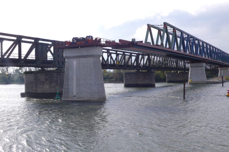 Das Bild zeigt eine große Brücke mit Stahlfachwerkkonstruktion, die über ein fließendes Gewässer führt und von mehreren Betonpfeilern getragen wird. Im Wasser nahe der Pfeiler sind Bojen zu sehen, der Himmel ist teils bewölkt.