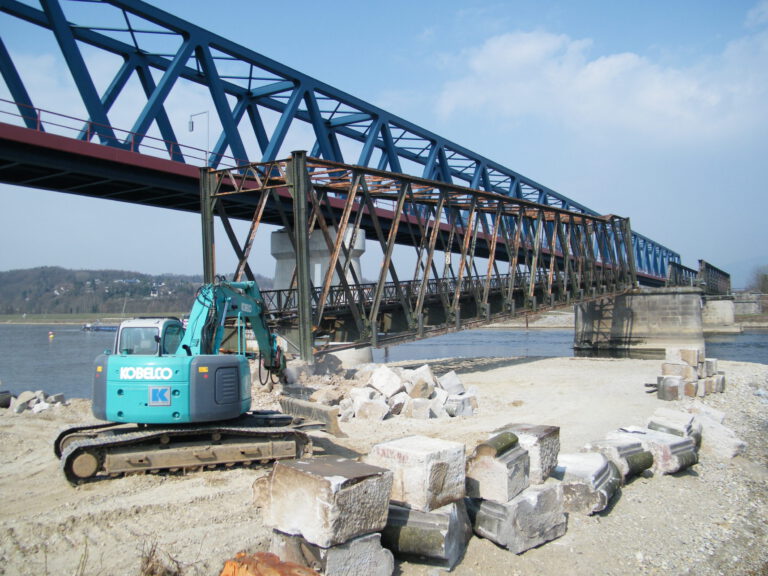 Das Bild zeigt eine Baustelle an einem Fluss mit einem großen blauen Stahlfachwerkbrücke im Hintergrund, die von Betonpfeilern getragen wird. Links im Bild steht ein Bagger, umgeben von großen Steinblöcken.