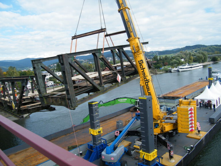 Das Bild zeigt einen großen gelben Kran, der ein Brückenelement über einem Fluss hebt. Weitere Maschinen und Hilfskonstruktionen sind zu sehen, im Hintergrund befinden sich Gebäude, Boote, Bäume und Hügel.