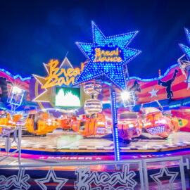 Ein farbenfrohes Fahrgeschäft namens ‚Break Dance‘ mit orange-gelben Wagen dreht sich auf einer runden Plattform. Im Hintergrund sind bunte Lichter, Sterne und Tänzer-Silhouetten zu sehen.
