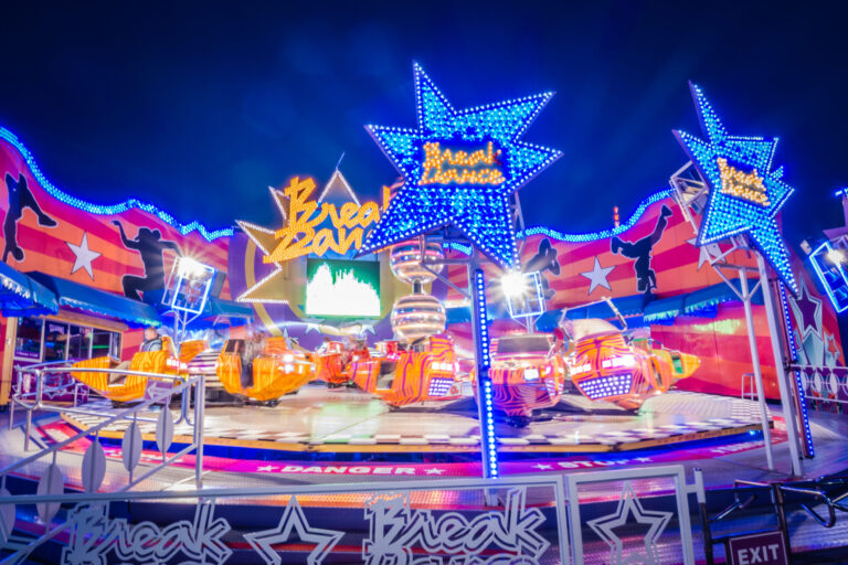Ein farbenfrohes Fahrgeschäft namens ‚Break Dance‘ mit orange-gelben Wagen dreht sich auf einer runden Plattform. Im Hintergrund sind bunte Lichter, Sterne und Tänzer-Silhouetten zu sehen.