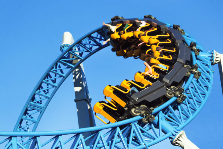Eine Achterbahn mit gelben und schwarzen Wagen fährt durch eine Looping-Kurve auf einer blauen Schiene. Der Himmel im Hintergrund ist klar und blau.