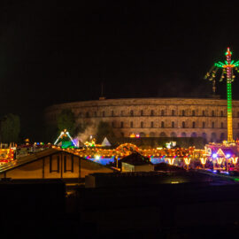 Das Nürnberger Volksfest bei Nacht mit beleuchtetem Riesenrad, Achterbahn mit gelben Schienen und weiteren Fahrgeschäften. Im Hintergrund ist das Kolosseum zu erkennen.