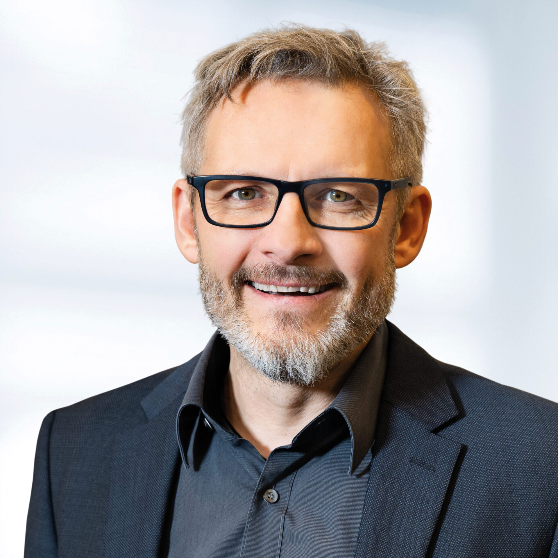 Edgar Görl trägt ein dunkles Sakko und ein dunkles Hemd. Der Hintergrund ist unscharf und zeigt ein Büro.