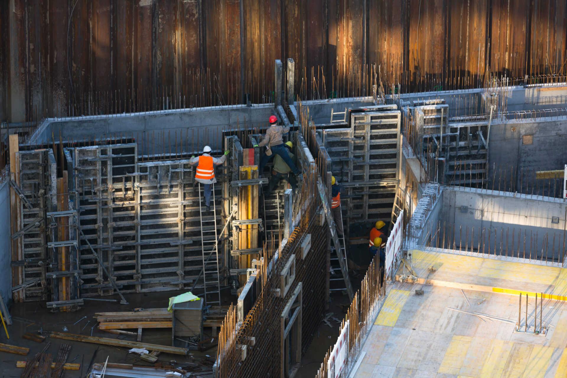 Das Bild zeigt eine Baustelle mit mehreren Arbeitern, die Schutzkleidung wie Helme und Warnwesten tragen. Die Arbeiter sind mit dem Bau einer Betonstruktur beschäftigt, umgeben von Metallgerüsten und Holzbohlen. Auf dem Gelände liegen verschiedene Baumaterialien, und die Baustelle ist von hohen Metallwänden eingefasst.