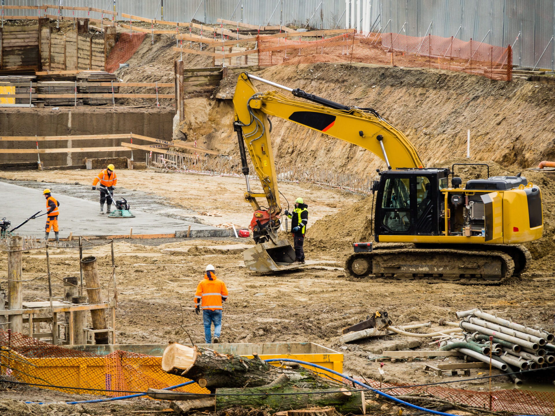 Das Bild zeigt eine Baustelle mit mehreren Arbeitern und Baumaschinen. Im Vordergrund befindet sich ein großer gelber Bagger, der von einem Arbeiter mit Schutzkleidung bedient wird. Weitere Arbeiter sind im Hintergrund zu sehen, einer davon arbeitet mit einem Elektrowerkzeug an einer Betonfläche. Auf dem Gelände liegen verschiedene Baumaterialien wie Holzträger und Metallrohre, die Baustelle ist mit provisorischen Zäunen und Absperrungen gesichert.