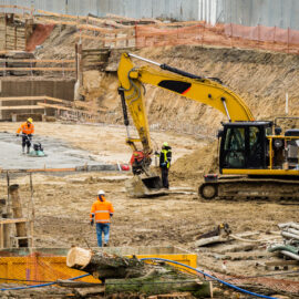 Das Bild zeigt eine Baustelle mit mehreren Arbeitern und Baumaschinen. Im Vordergrund befindet sich ein großer gelber Bagger, der von einem Arbeiter mit Schutzkleidung bedient wird. Weitere Arbeiter sind im Hintergrund zu sehen, einer davon arbeitet mit einem Elektrowerkzeug an einer Betonfläche. Auf dem Gelände liegen verschiedene Baumaterialien wie Holzträger und Metallrohre, die Baustelle ist mit provisorischen Zäunen und Absperrungen gesichert.