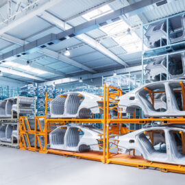 Das Bild zeigt ein Industriegebäude mit mehreren gestapelten Karosserierahmen, die auf Metallgestellen gelagert sind. Einige der Gestelle sind orange lackiert, andere haben eine metallische Farbe. Die Halle hat eine hohe Decke mit Oberlichtern und Hängelampen zur Beleuchtung.