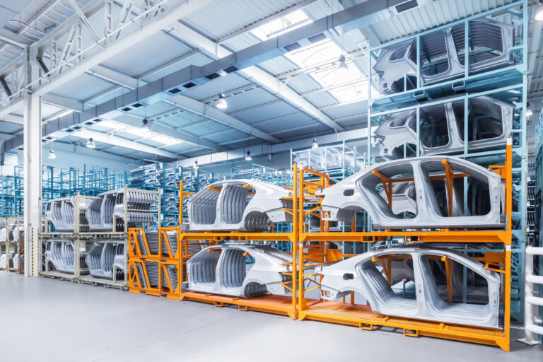 Das Bild zeigt ein Industriegebäude mit mehreren gestapelten Karosserierahmen, die auf Metallgestellen gelagert sind. Einige der Gestelle sind orange lackiert, andere haben eine metallische Farbe. Die Halle hat eine hohe Decke mit Oberlichtern und Hängelampen zur Beleuchtung.