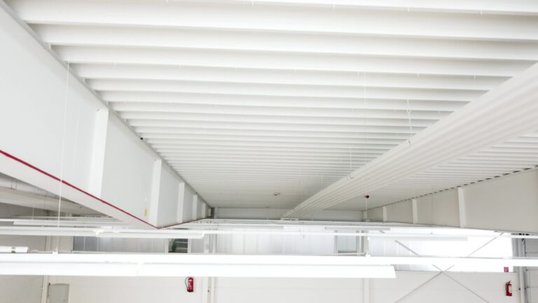 Das Bild zeigt die Decke eines Industrie- oder Gewerbegebäudes, bestehend aus weißen Metallträgern und -paneelen. Mehrere Leuchtstofflampen hängen von der Decke, und ein rotes Rohr des Sprinklersystems verläuft waagrecht entlang der Decke. An der Wand darunter ist ein Feueralarm oder Feuerlöscher montiert.