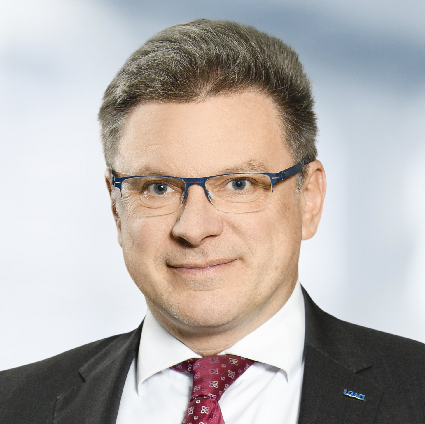 Michael Handrieder trägt einen Anzug mit weißem Hemd und roter Krawatte. Der Hintergrund ist unscharf und zeigt ein Büro.