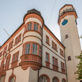 Historisches Rathaus in Hof mit mehreren Etagen, großen Fenstern und dekorativen Elementen in Rot und Weiß, ergänzt durch einen angebauten Uhrturm mit blauer Uhr; rechts im Bild sind teilweise Bäume zu sehen.