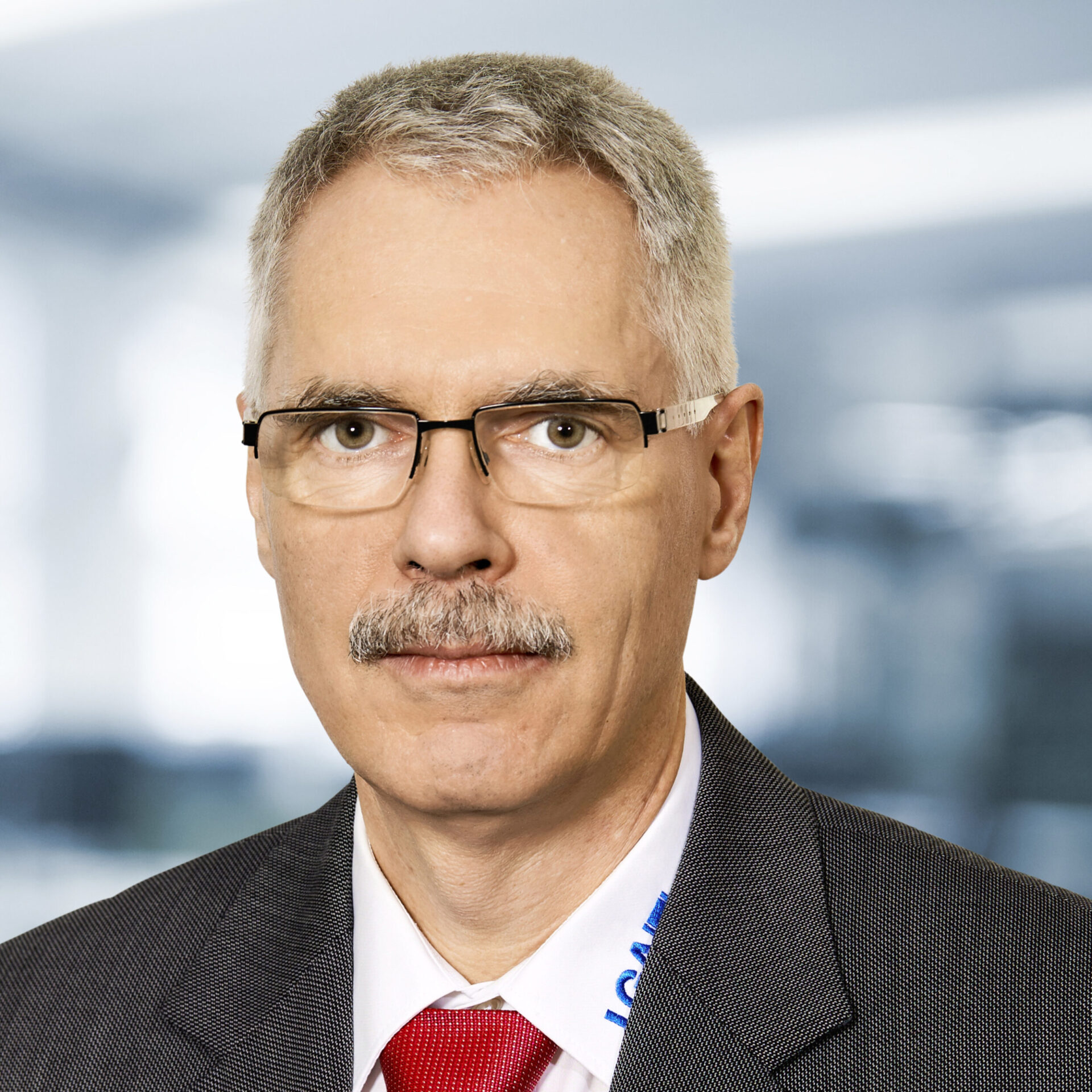 Peter Hofmann trägt einen Anzug mit weißem Hemd und roter Krawatte. Der Hintergrund ist unscharf und zeigt ein Büro.