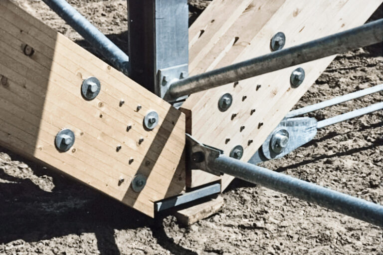 Das Bild zeigt eine Nahaufnahme eines Holzknotenpunkts auf einer Baustelle, bei dem mehrere Holzbalken mit Metallplatten und Bolzen verbunden sind. Zusätzliche Metallstreben sorgen für Stabilität, der Untergrund besteht aus Erde.