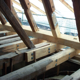 Das Bild zeigt eine hölzerne Dachkonstruktion im Bau, bestehend aus mehreren Balken und Sparren, die mit Metallplatten und Bolzen verbunden sind. Im Hintergrund ist ein Sicherheitsnetz gespannt.