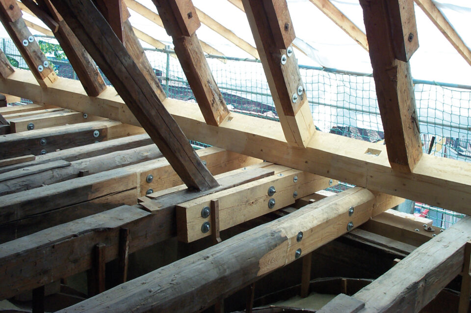 Das Bild zeigt eine hölzerne Dachkonstruktion im Bau, bestehend aus mehreren Balken und Sparren, die mit Metallplatten und Bolzen verbunden sind. Im Hintergrund ist ein Sicherheitsnetz gespannt.