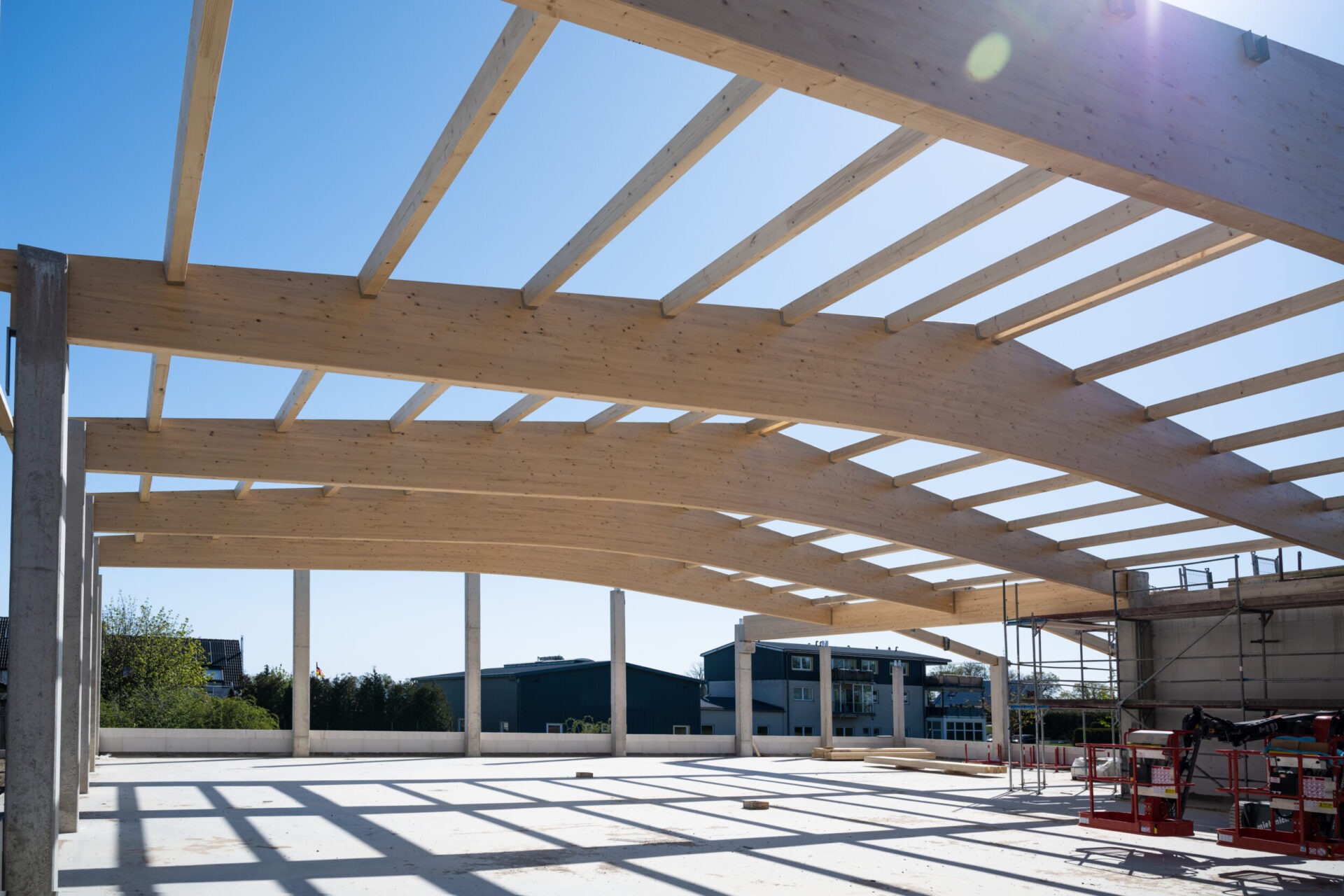 Das Bild zeigt den Rohbau einer Dachkonstruktion mit gebogenen Holzbalken, die von senkrechten Betonsäulen getragen werden. Auf der rechten Seite sind Gerüste und Baumaschinen zu sehen. Im Hintergrund befinden sich Gebäude und Bäume unter einem klaren blauen Himmel.