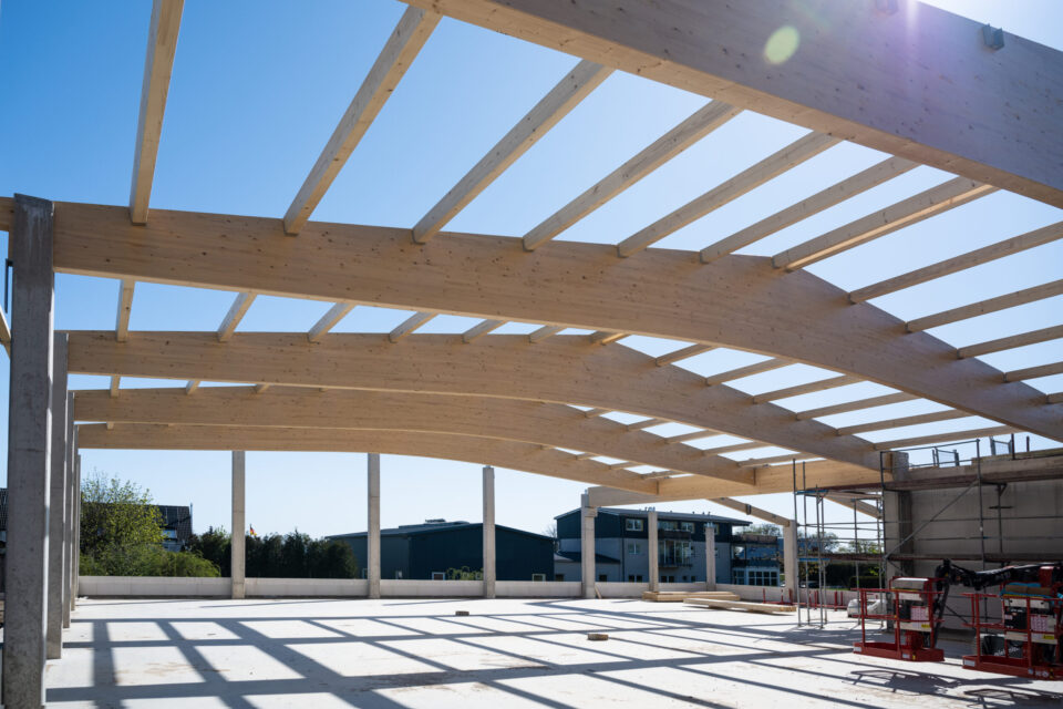 Das Bild zeigt den Rohbau einer Dachkonstruktion mit gebogenen Holzbalken, die von senkrechten Betonsäulen getragen werden. Auf der rechten Seite sind Gerüste und Baumaschinen zu sehen. Im Hintergrund befinden sich Gebäude und Bäume unter einem klaren blauen Himmel.