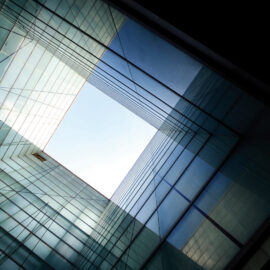 Das Bild zeigt eine moderne Architektur mit Glaswänden aus der Froschperspektive. Die Anordnung der Glasflächen bildet ein geometrisches Muster, das den Blick auf eine quadratische Öffnung im Himmel lenkt. Lichtreflexionen erzeugen unterschiedliche Blau- und Grüntöne, die dem Bild Tiefe verleihen.