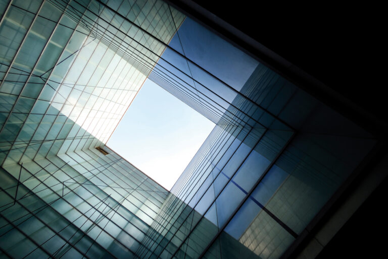 Das Bild zeigt eine moderne Architektur mit Glaswänden aus der Froschperspektive. Die Anordnung der Glasflächen bildet ein geometrisches Muster, das den Blick auf eine quadratische Öffnung im Himmel lenkt. Lichtreflexionen erzeugen unterschiedliche Blau- und Grüntöne, die dem Bild Tiefe verleihen.