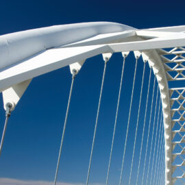 Das Bild zeigt eine moderne weiße Hängebrücke mit mehreren senkrechten Seilen, die an einer bogenförmigen Struktur befestigt sind. Die Konstruktion weist geometrische Muster und dreieckige Stützelemente auf, der Himmel ist klar und blau.