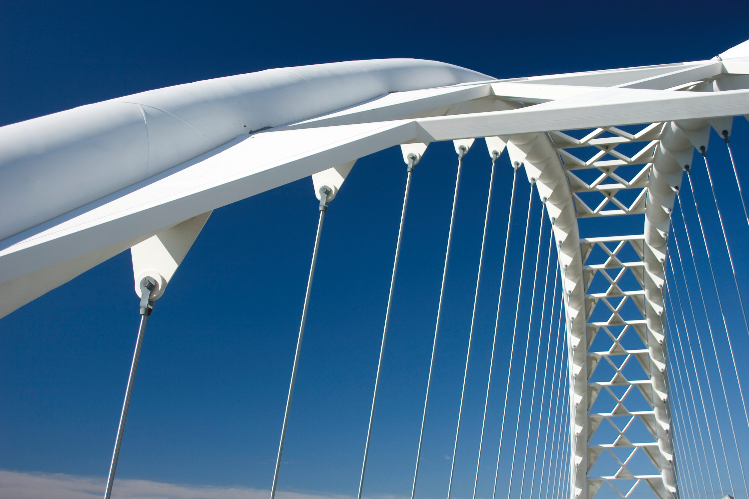 Das Bild zeigt eine moderne weiße Hängebrücke mit mehreren senkrechten Seilen, die an einer bogenförmigen Struktur befestigt sind. Die Konstruktion weist geometrische Muster und dreieckige Stützelemente auf, der Himmel ist klar und blau.